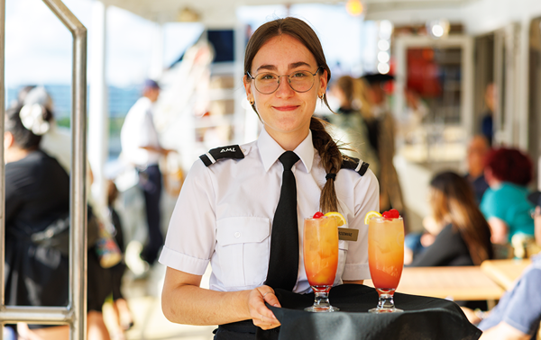 Une serveuse souriante en uniforme tenant un plateau avec deux cocktails de couleur orangée posés dessus, le tout sur le navire AML Cavalier Maxim à Montréal. 