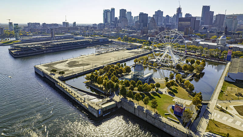 Vue aérienne d'un grand parc urbain entouré d'eau, avec une grande roue, des espaces verts et des bâtiments modernes en arrière-plan dans une ville.