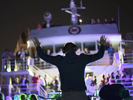 Un DJ anime une fête nocturne sur un bateau, avec des lumières colorées et une foule dansante sur plusieurs niveaux du navire.