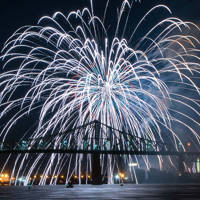 A spectacular firework display bursts in the night sky above an illuminated bridge, with reflections on the water and city lights in the background.