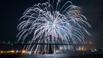 De magnifiques feux d'artifice blancs éclatent dans le ciel nocturne au-dessus d'un pont illuminé, avec des reflets scintillants sur l'eau et les lumières de la ville en arrière-plan.