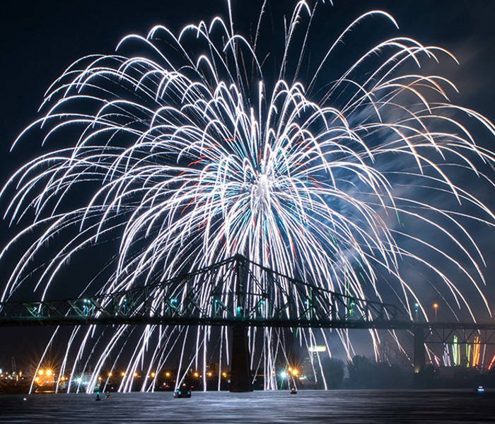 De magnifiques feux d'artifice blancs éclatent dans le ciel nocturne au-dessus d'un pont illuminé, avec des reflets scintillants sur l'eau et les lumières de la ville en arrière-plan.