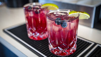 Deux verres d'un cocktail rouge vif reposent sur un tapis de bar. Chaque boisson est ornée d'une tranche de lime et de quelques myrtilles. Les verres, au design raffiné, contiennent des glaçons. L'ensemble est posé sur le comptoir d'un bar ou d'un restaurant.