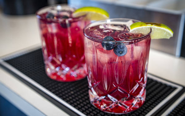 Deux verres d'un cocktail rouge vif reposent sur un tapis de bar. Chaque boisson est ornée d'une tranche de lime et de quelques myrtilles. Les verres, au design raffiné, contiennent des glaçons. L'ensemble est posé sur le comptoir d'un bar ou d'un restaurant.