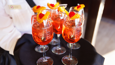 A close-up of a server's tray with 6 glasses of orange-colored Spritz, each garnished with an orange wedge and a cherry.