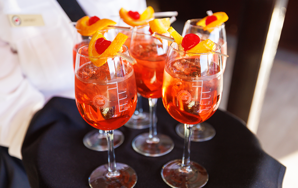 A close-up of a server's tray with 6 glasses of orange-colored Spritz, each garnished with an orange wedge and a cherry.