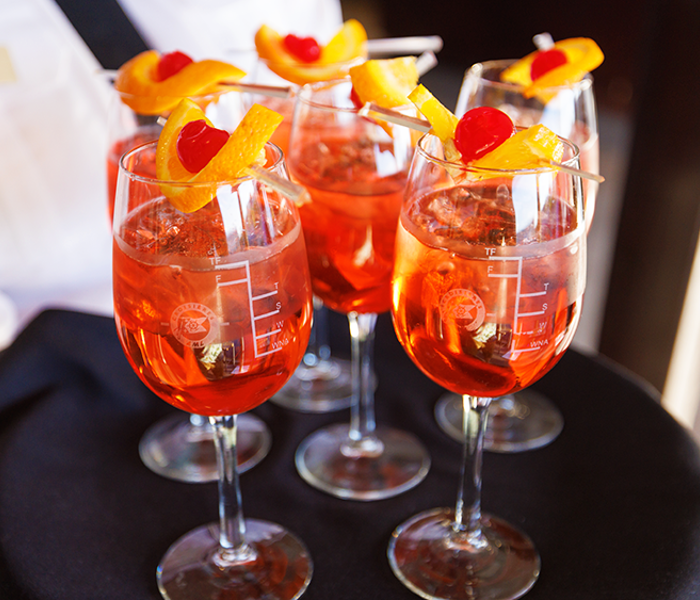 A close-up of a server's tray with 6 glasses of orange-colored Spritz, each garnished with an orange wedge and a cherry.