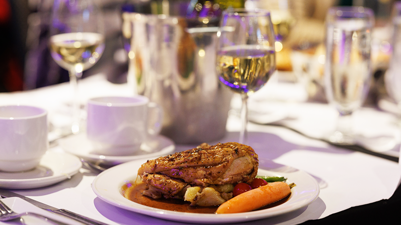 A bistro-style plate featuring a chicken supreme, baby potatoes, and fresh market vegetables, accompanied by a glass of white wine.