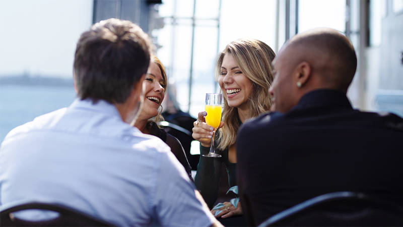 Quatre personnes sont assises ensemble à une table en extérieur, riant et profitant de leurs boissons. Une femme aux cheveux longs tient un verre de jus d'orange. L'endroit semble ensoleillé et offre une vue sur l'eau.