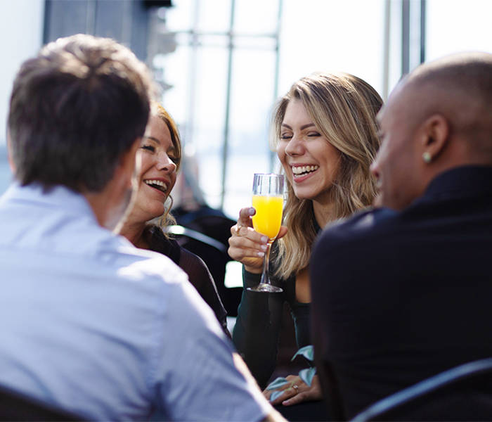 Quatre personnes sont assises ensemble à une table en extérieur, riant et profitant de leurs boissons. Une femme aux cheveux longs tient un verre de jus d'orange. L'endroit semble ensoleillé et offre une vue sur l'eau.