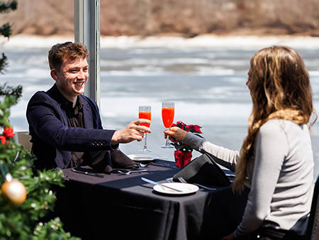  Un couple trinque avec des cocktails rouges lors d'un repas élégant près d'une fenêtre avec vue sur une rivière, avec un sapin de Noël décoré en arrière-plan.