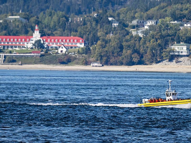 A yellow boat with several passengers speeds across a wide blue river. In the background, a building with a red roof and a lighthouse tower is situated on a tree-lined shore.