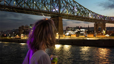 Une femme de dos tenant un verre, admirant un pont illuminé traversant une rivière, avec des lumières urbaines et un ciel au crépuscule en arrière-plan.