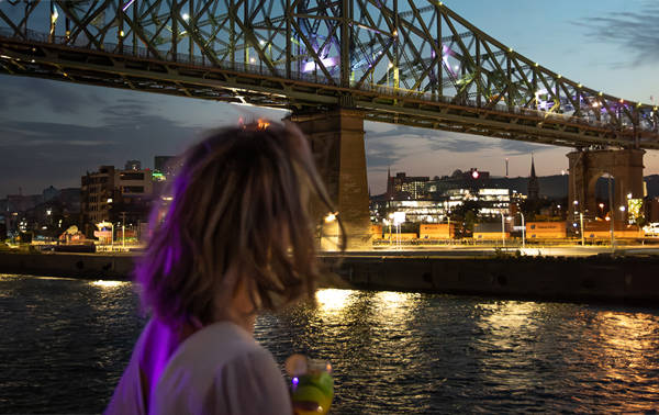 Une femme de dos tenant un verre, admirant un pont illuminé traversant une rivière, avec des lumières urbaines et un ciel au crépuscule en arrière-plan.