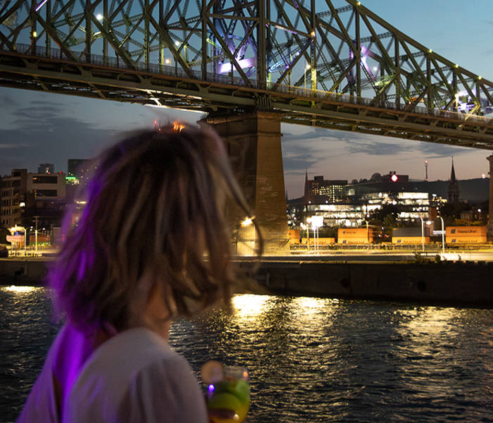 Une femme de dos tenant un verre, admirant un pont illuminé traversant une rivière, avec des lumières urbaines et un ciel au crépuscule en arrière-plan.