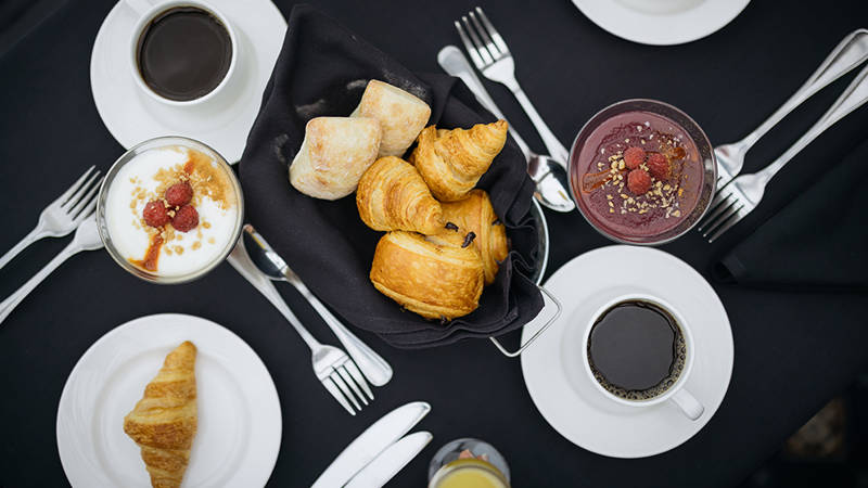 Vue aérienne d'une table de petit-déjeuner comprenant des croissants, des petits pains dans un panier, des tasses de café, un verre de smoothie garni de granola et de baies, ainsi qu'un parfait au yaourt et aux framboises. Les couverts sont soigneusement disposés autour des plats.