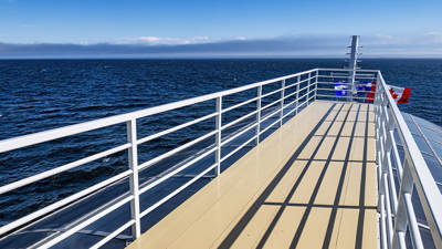 Vue depuis le pont d'un navire, où des balustrades blanches projettent leurs ombres sur le pont beige. L'immense océan s'étend sous un ciel bleu clair, avec un horizon lointain visible. Des bouées de sauvetage rouges et bleues sont fixées aux balustrades.