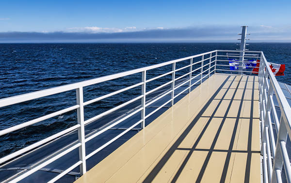 Vue depuis le pont d'un navire, où des balustrades blanches projettent leurs ombres sur le pont beige. L'immense océan s'étend sous un ciel bleu clair, avec un horizon lointain visible. Des bouées de sauvetage rouges et bleues sont fixées aux balustrades.