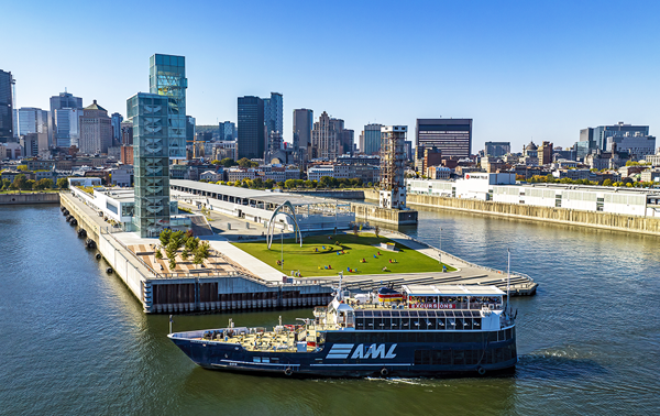 Vue aérienne du navire AML Cavalier Maxim stationné devant la Tour du Port de Montréal de jour avec vue sur la skyline ensoleillée de la ville en arrière plan.