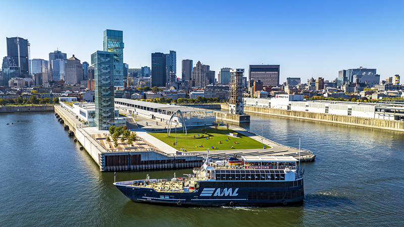 Aerial view of the AML Cavalier Maxim parked in front of the Port of Montreal Tower by day, with a view of the city's sunny skyline in the background.