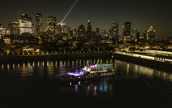 Vue aérienne du navire AML Cavalier Maxim stationné dans le Vieux-Port de Montréal de nuit avec vue sur la skyline de la ville en arrière plan.