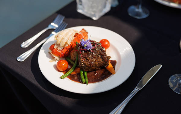 Un plat gastronomique servi sur une assiette blanche, comprenant un filet de viande nappé de sauce, des légumes, une queue de homard et une garniture florale, présenté sur une table élégante avec une nappe noire.