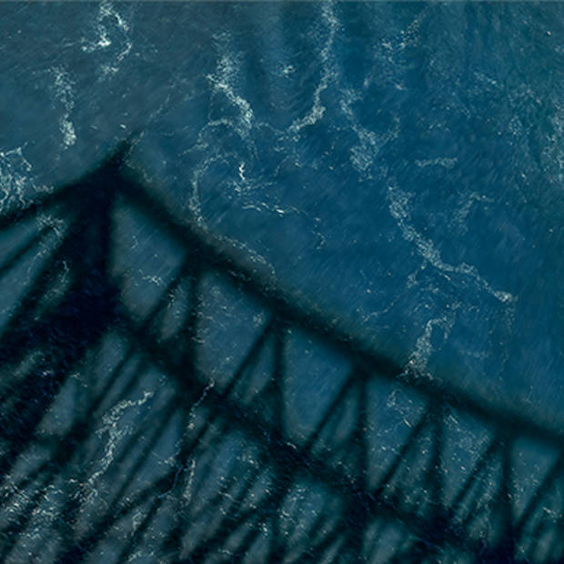 Aerial view of a boat sailing through the blue waters of the Saint Lawrence under a large bridge casting a shadow. The shadow creates a striking pattern on the surface of the water.