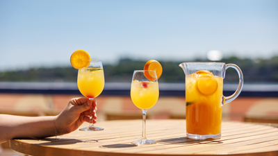 A close-up of a hand holding a glass of orange sangria, with another glass and a pitcher of sangria served on a wooden table on one of the terraces of the AML Cavalier Maxim in Montreal.