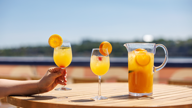 A close-up of a hand holding a glass of orange sangria, with another glass and a pitcher of sangria served on a wooden table on one of the terraces of the AML Cavalier Maxim in Montreal.