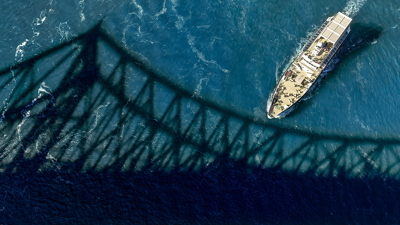 Aerial view of the Cavalier Maxime sailing on the sparkling St. Lawrence River, with the shadow of the Jacques Cartier Bridge in the foreground.