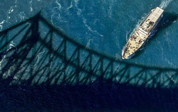 Vue aérienne du navire Cavalier Maxime naviguant sur l'étincelant fleuve Saint-Laurent avec l'ombre du Pont Jacques Cartier au premier plan. 