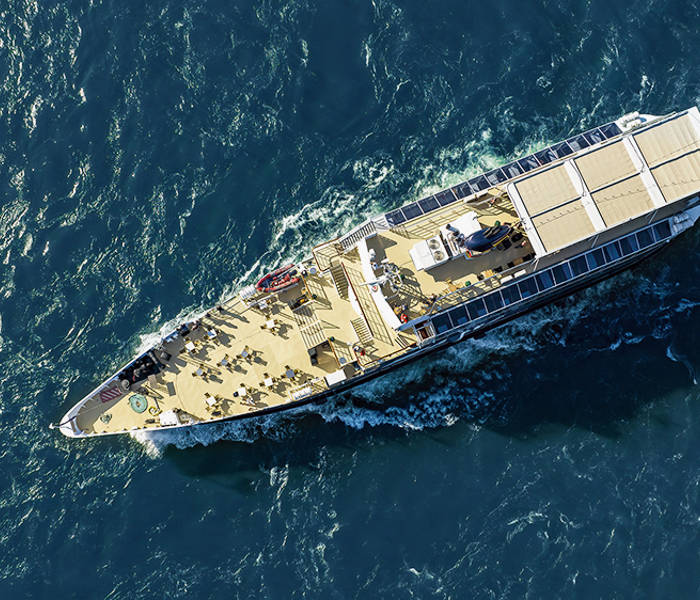 Vue aérienne d'un bateau de croisière naviguant sur des eaux bleues, avec des passagers sur le pont profitant du voyage.