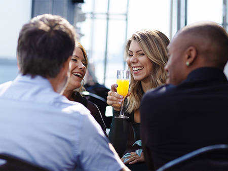 Un groupe de quatre amis partage un moment joyeux dans un restaurant près de l'eau, une femme souriante tient un verre de mimosa.