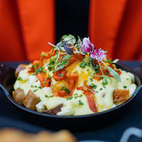 A delicious dish of roasted potatoes topped with hollandaise sauce, garnished with vegetables, fresh herbs, and edible flowers, served in a cast-iron skillet.