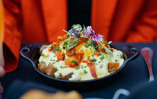 Un plat gourmand composé de pommes de terre rôties, nappées de sauce hollandaise, garnies de légumes, herbes fraîches et fleurs comestibles, servi dans une poêle en fonte.