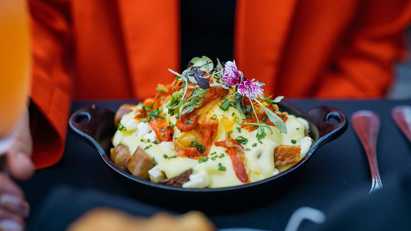 Un plat gourmand composé de pommes de terre rôties, nappées de sauce hollandaise, garnies de légumes, herbes fraîches et fleurs comestibles, servi dans une poêle en fonte.