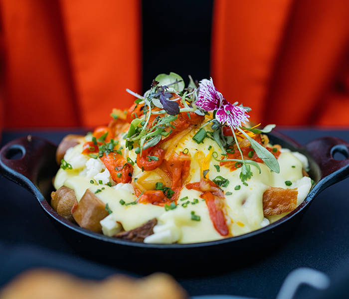 Un plat gourmand composé de pommes de terre rôties, nappées de sauce hollandaise, garnies de légumes, herbes fraîches et fleurs comestibles, servi dans une poêle en fonte.