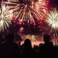 Une foule de spectateurs regarde un spectaculaire feu d'artifice illuminant le ciel nocturne avec des éclats dorés et rouges. La scène est empreinte d'émerveillement et de célébration.