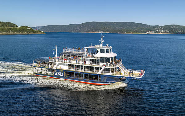 Un grand ferry portant les lettres "AML" navigue sur une étendue d'eau. C'est une journée ensoleillée avec un ciel dégagé. À l'arrière-plan, on aperçoit des collines verdoyantes et une petite ville avec des bâtiments aux toits rouges le long du rivage.