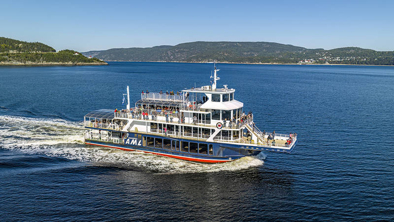 Un grand ferry portant les lettres "AML" navigue sur une étendue d'eau. C'est une journée ensoleillée avec un ciel dégagé. À l'arrière-plan, on aperçoit des collines verdoyantes et une petite ville avec des bâtiments aux toits rouges le long du rivage.