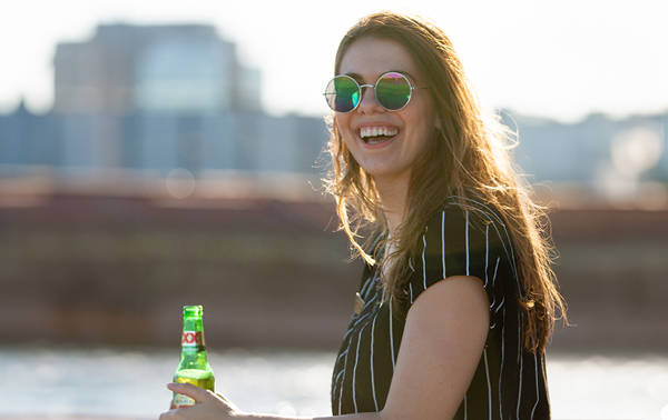 Une femme souriante portant des lunettes de soleil rondes et une chemise à rayures, tenant une bouteille de bière verte, avec une rivière et des bâtiments floutés en arrière-plan.