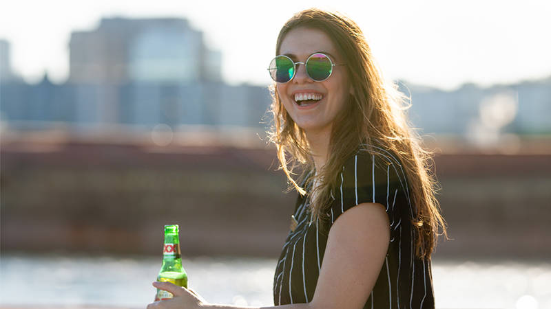 Une femme souriante portant des lunettes de soleil rondes et une chemise à rayures, tenant une bouteille de bière verte, avec une rivière et des bâtiments floutés en arrière-plan.