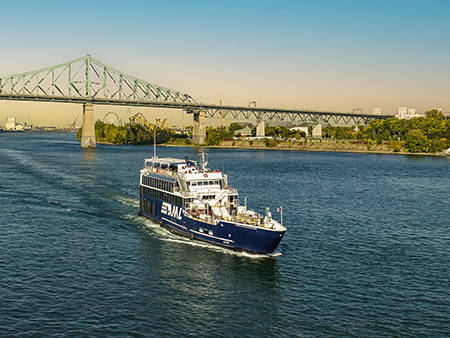 Un bateau de croisière bleu navigue sur une rivière par une journée ensoleillée, avec le pont Jacques-Cartier et des arbres en arrière-plan.