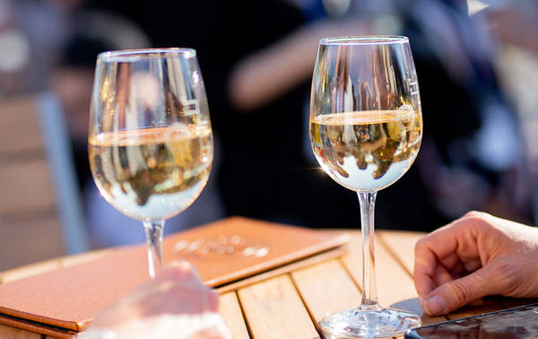 Deux verres de vin blanc posés sur une table en bois, avec des menus en cuir et une main visible au premier plan. L'arrière-plan est flou, suggérant une ambiance extérieure conviviale et lumineuse.