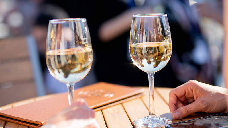 Deux verres de vin blanc posés sur une table en bois, avec des menus en cuir et une main visible au premier plan. L'arrière-plan est flou, suggérant une ambiance extérieure conviviale et lumineuse.