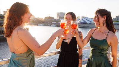 Trois femmes élégamment habillées trinquent avec des cocktails orangés sur le pont d'un bateau, avec une vue sur l'eau et la ville sous un coucher de soleil.