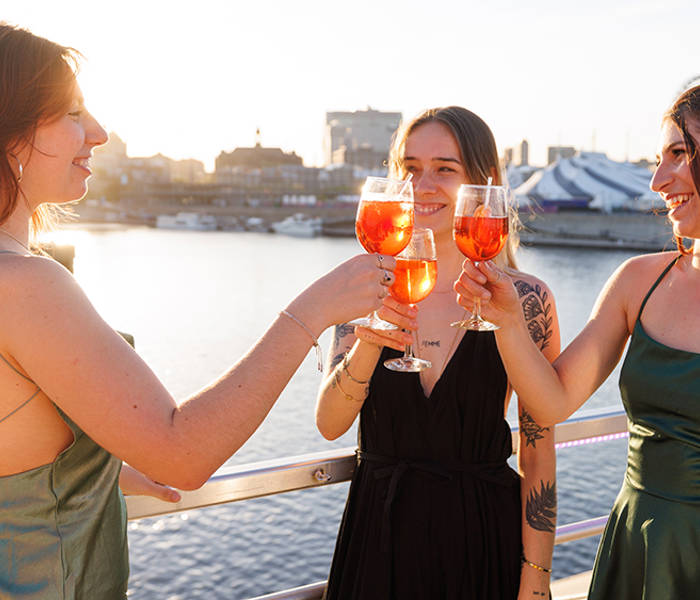 Trois femmes élégamment habillées trinquent avec des cocktails orangés sur le pont d'un bateau, avec une vue sur l'eau et la ville sous un coucher de soleil.