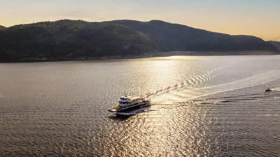 Un ferry navigue sur une étendue d'eau paisible sous un coucher de soleil doré. Le ciel est partiellement nuageux, et des collines recouvertes d'une dense forêt verte encadrent la scène en arrière-plan. Le ferry laisse derrière lui un sillage doux qui se dissipe lentement.