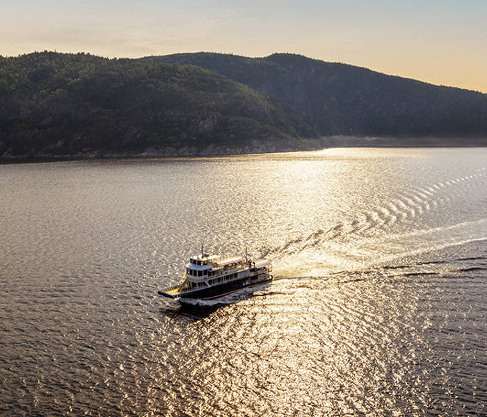 Un ferry navigue sur une étendue d'eau paisible sous un coucher de soleil doré. Le ciel est partiellement nuageux, et des collines recouvertes d'une dense forêt verte encadrent la scène en arrière-plan. Le ferry laisse derrière lui un sillage doux qui se dissipe lentement.