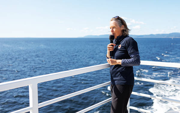 Une naturaliste se tient sur le pont d'un bateau, tenant un microphone, face à l'immensité de l'océan sous un ciel bleu clair. Des formations terrestres lointaines sont visibles à travers l'eau. La personne porte une tenue sombre et semble parler ou chanter.